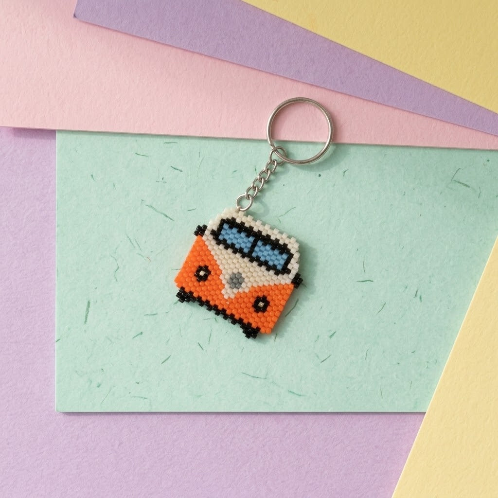 A small beaded keychain shaped like a retro orange and white van with blue windows, attached to a silver metal ring, displayed on pastel paper sheets.