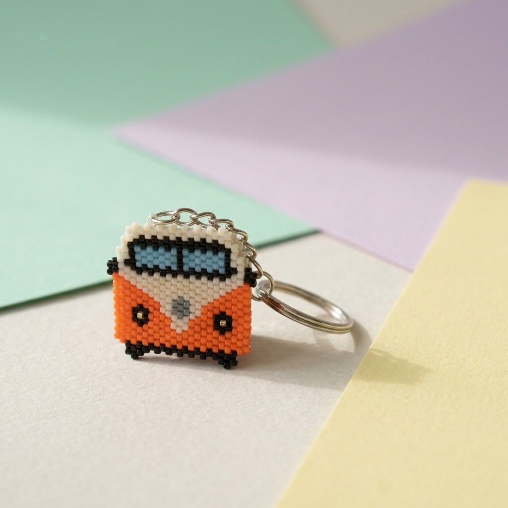 A beaded keychain shaped like a retro orange and white bus with a silver metal key ring, resting on a light beige surface with pastel green, purple, and yellow paper sheets in the background.