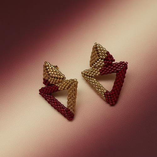 Pair of triangle-shaped earrings with gold and red beads on a pink background