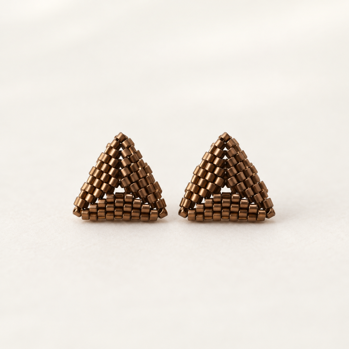 A pair of handmade triangular stud earrings crafted from metallic bronze seed beads, displayed on a clean, light off-white background.