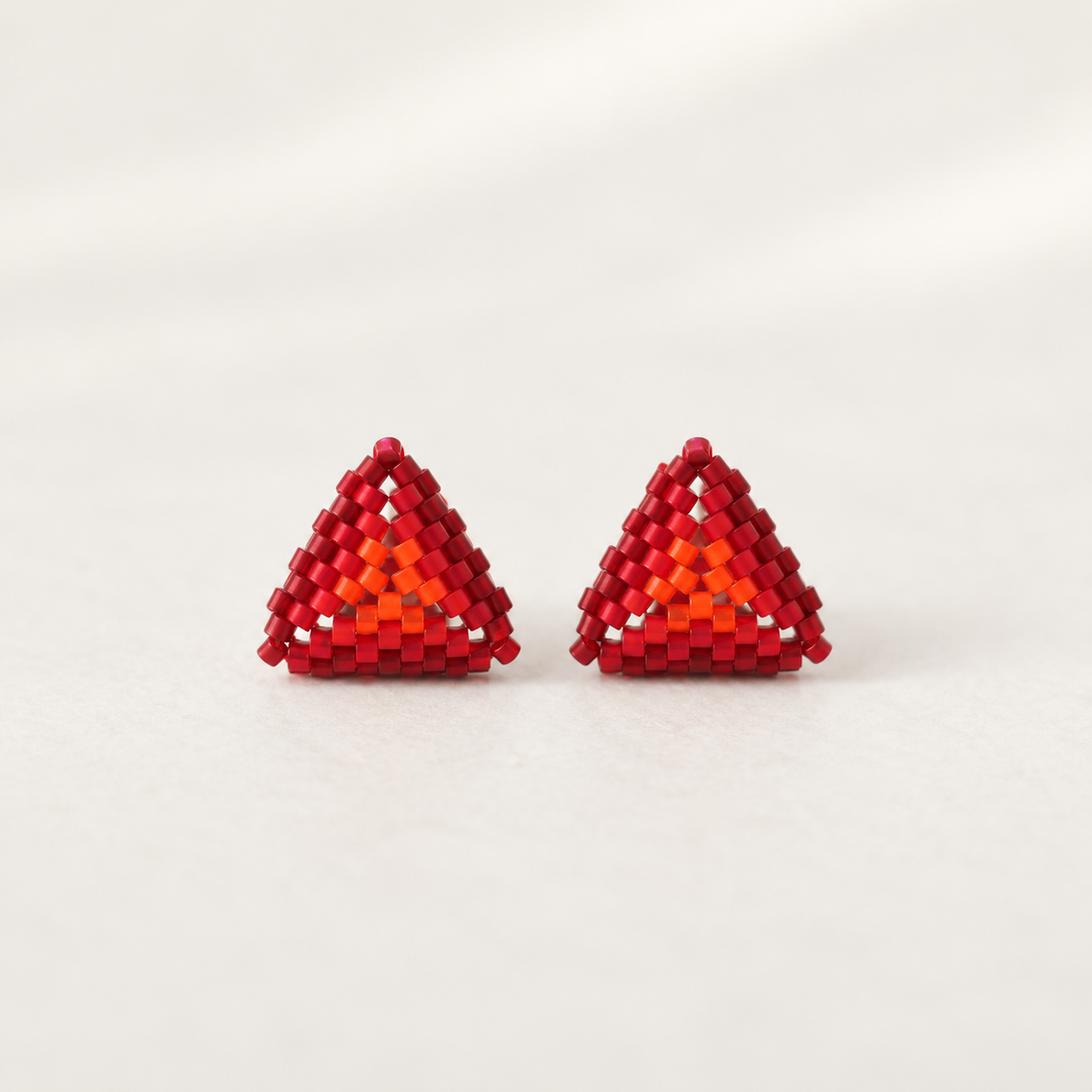 A pair of red triangular beaded earrings with a layered design featuring red, orange, and red beads, displayed on a light cream background.