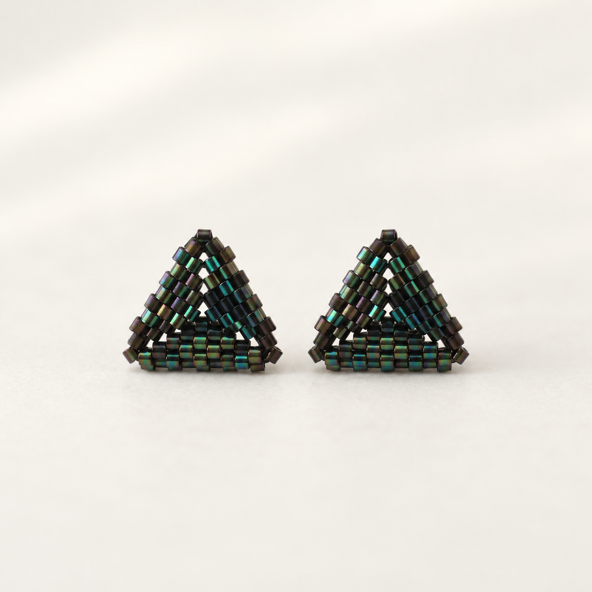A pair of handmade triangular beaded earrings made from small dark metallic beads with iridescent green and blue reflections, set against a plain light background.