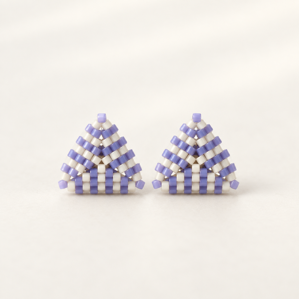 A pair of triangular beaded earrings made of white and light purple seed beads, arranged in a geometric pattern on a light cream background.