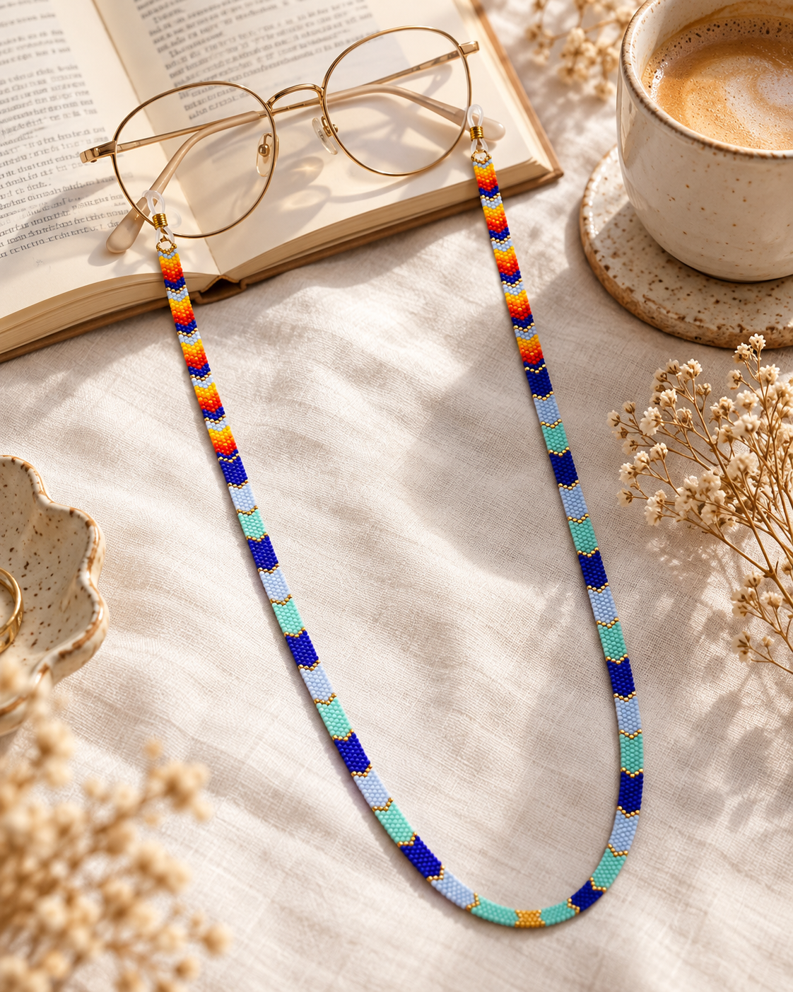 A colorful woven eyeglasses chain with blue, teal, yellow, orange, and red segments, shown hanging from gold-rimmed glasses placed on an open book, with a cup of coffee and dried flowers around it.