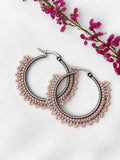 A pair of silver hoop earrings adorned with delicate rows of small pink beads, displayed on a textured white fabric background with a sprig of pink flowers in the corner.