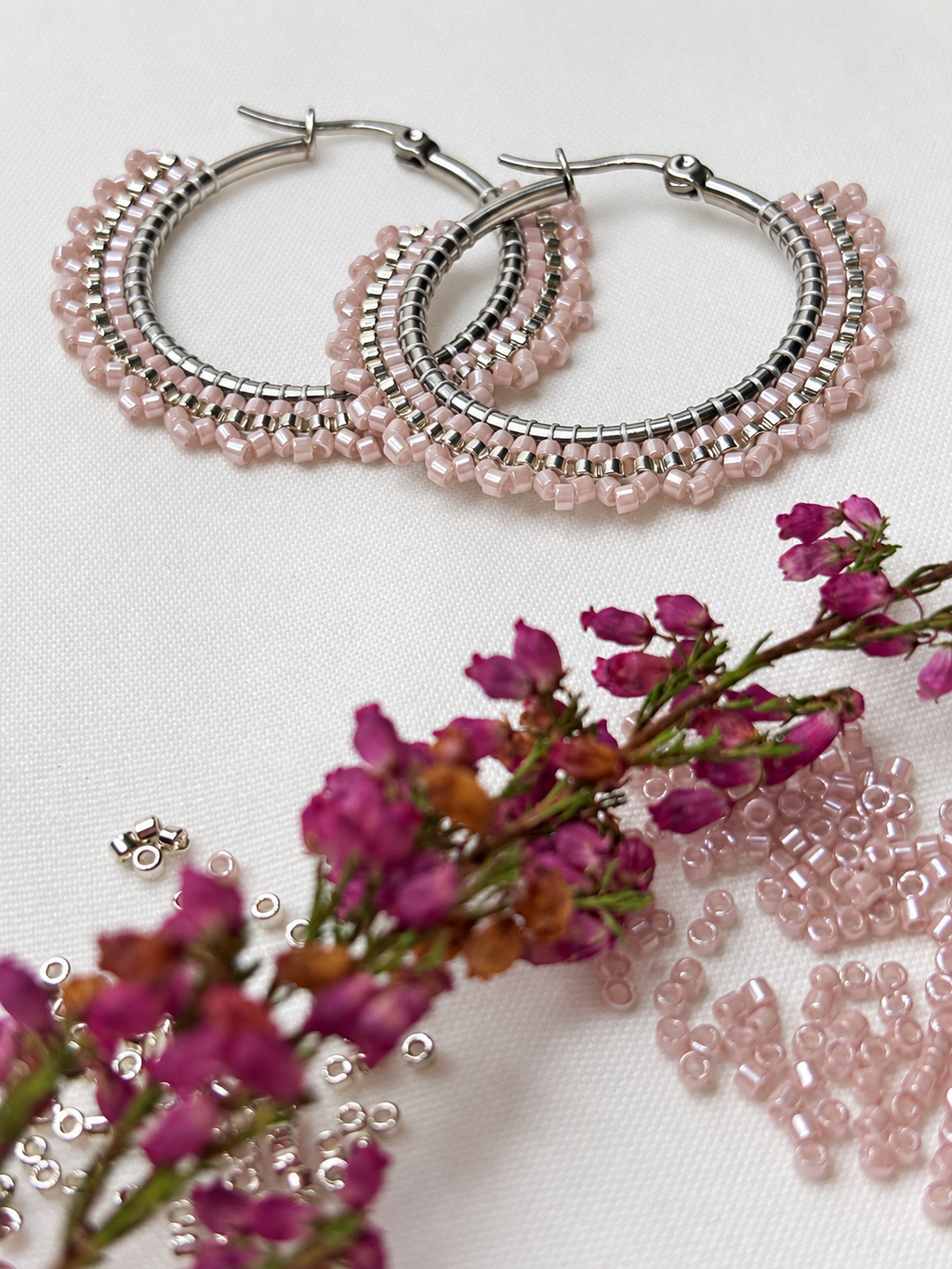 Silver-tone hoop earrings with multiple rows of small pink faceted beads, arranged on a white fabric background with pink flowers and loose beads as styling props.