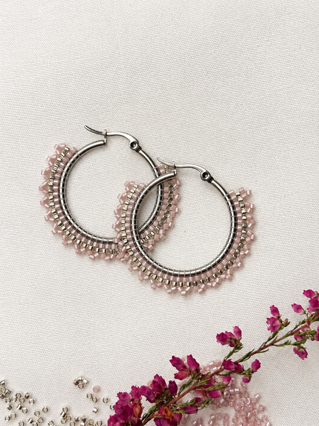 A pair of silver-tone hoop earrings with delicate rows of small light pink beads, arranged on a textured off-white fabric background with dried pink flowers and loose beads as styling props.