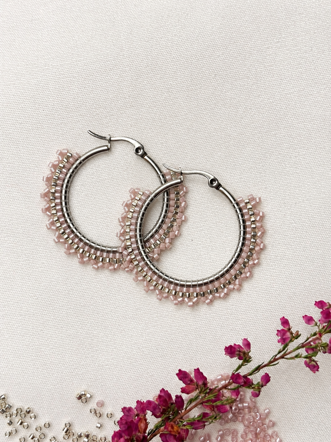 A pair of silver-tone hoop earrings with delicate rows of small light pink beads, arranged on a textured off-white fabric background with dried pink flowers and loose beads as styling props.
