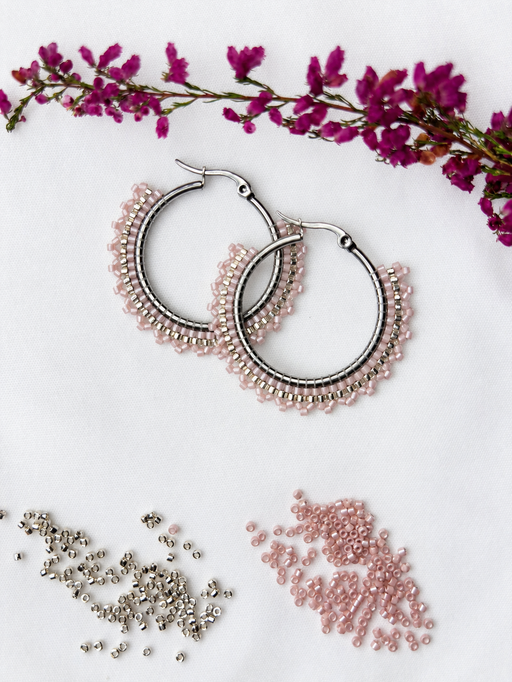 A pair of silver hoop earrings embellished with rows of small pale pink beads, arranged on a white background with scattered pink and silver beads and a sprig of purple flowers.