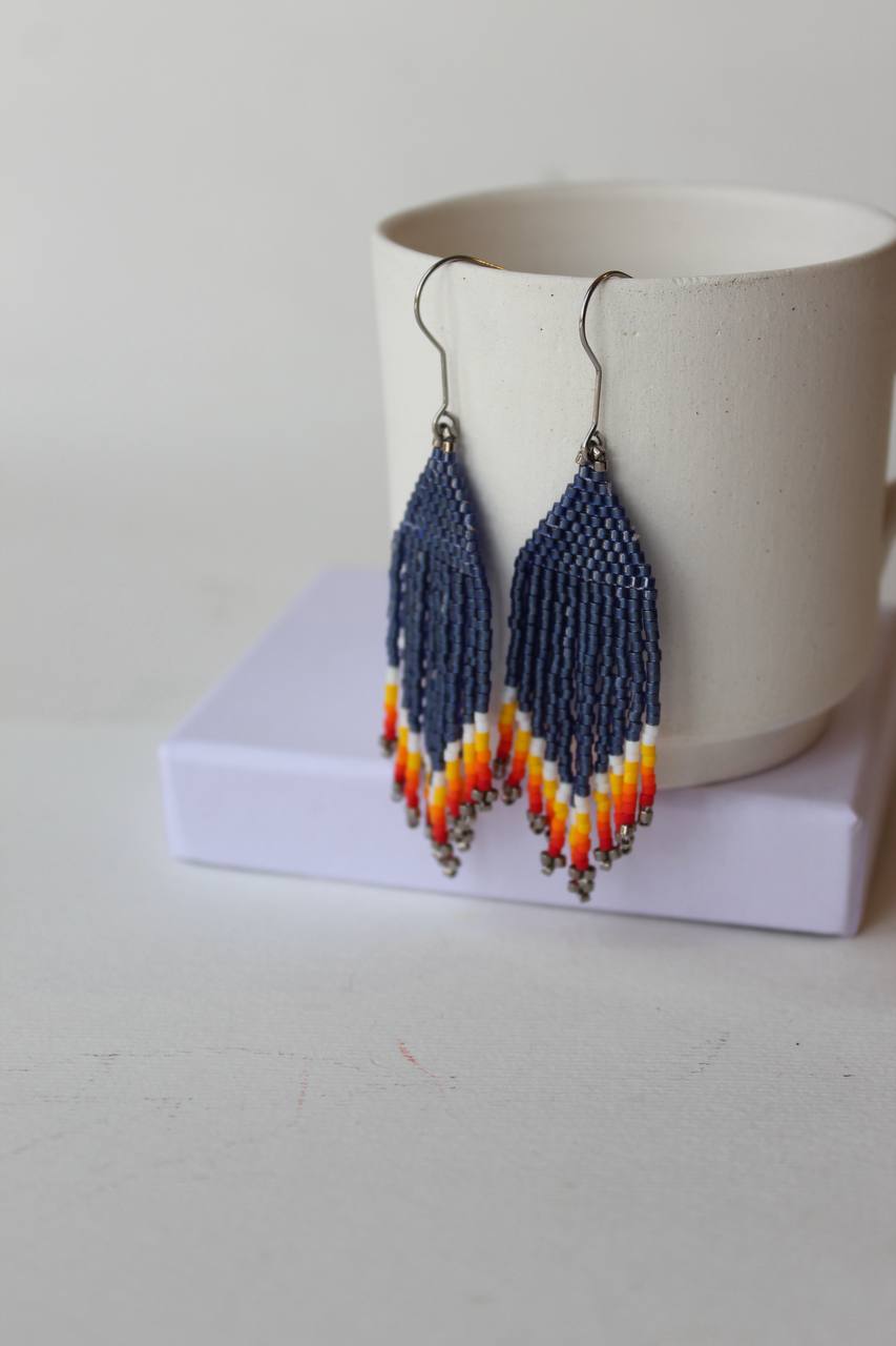 Ein Paar handgefertigte Perlenohrringe mit geometrischen Mustern in Marineblau, Orange, Gelb und Weiß, die an silbernen Angelhakendrähten an einer weißen Keramiktasse hängen.