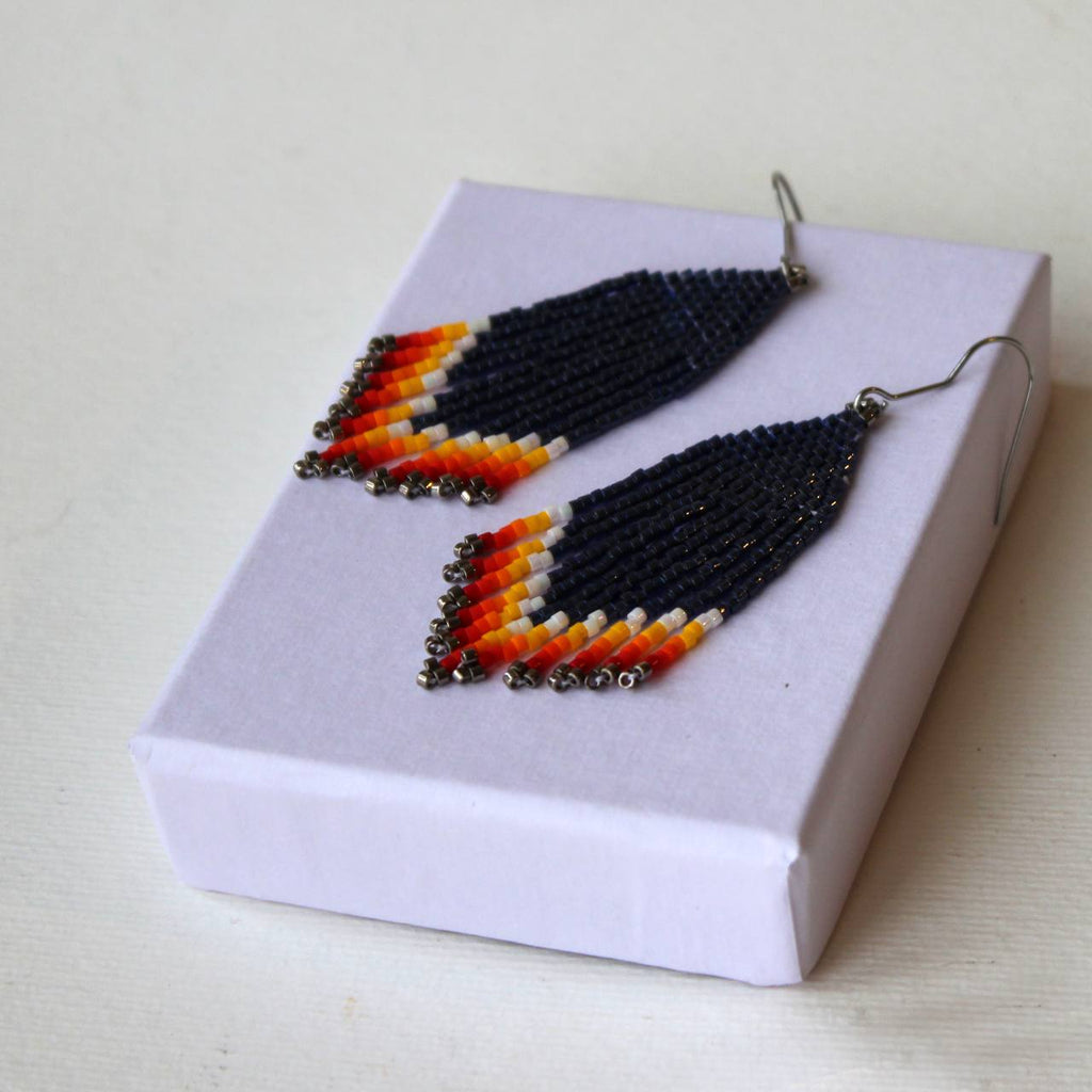 Ein Paar handgefertigte Perlenohrringe mit schwarzem Körper und mehrfarbigem Chevronmuster in Orange, Gelb und Weiß, präsentiert in einer Geschenkbox in hellem Lavendel.