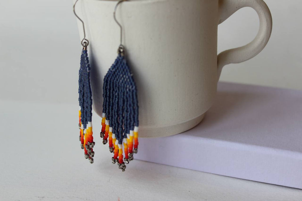 Ein Paar lange Perlenohrringe mit marineblauen, weißen, orangefarbenen und gelben Perlen, die vor einer weißen Keramiktasse auf einer hellen Oberfläche hängen.