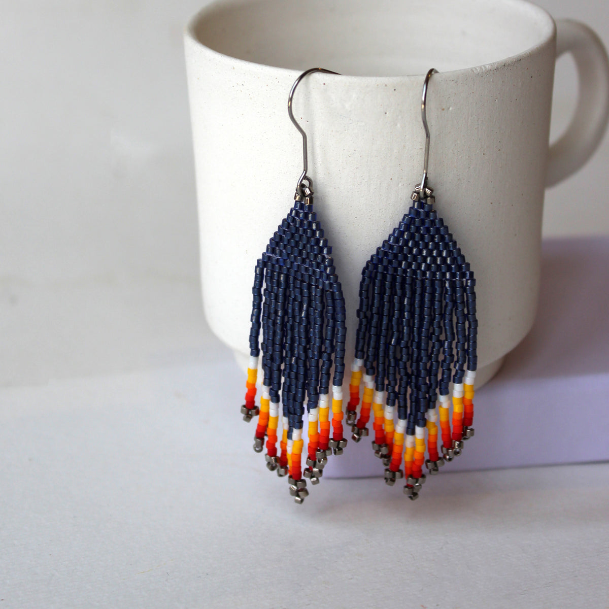 Ein Paar handgefertigte Perlenohrringe mit geometrischen Mustern in Marineblau, Orange, Gelb und Weiß, die vor einer weißen Keramiktasse hängen.