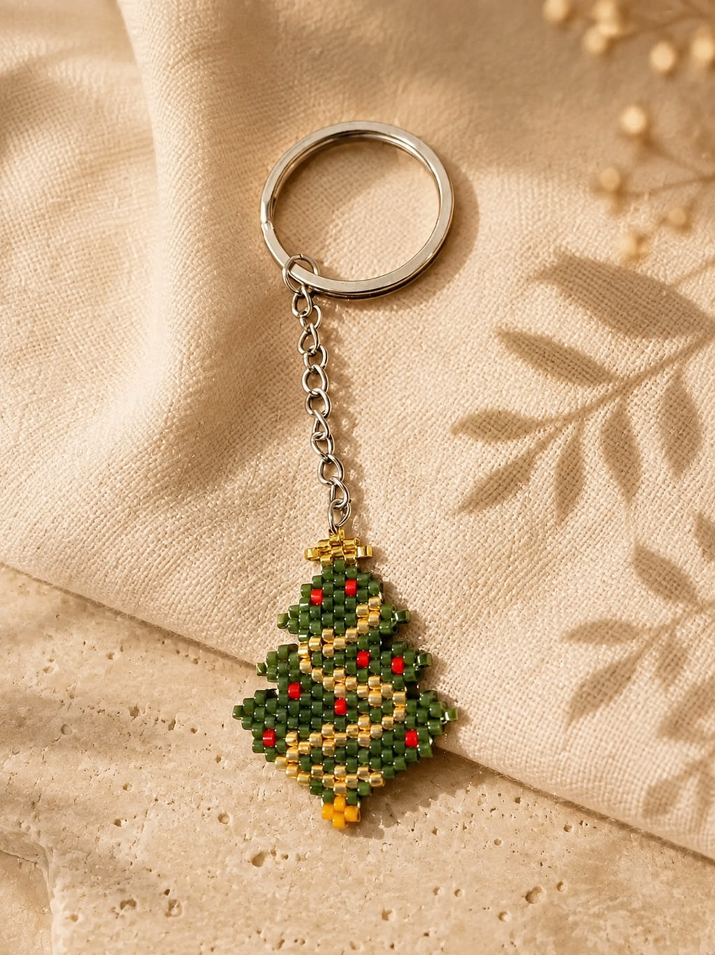 A beaded Christmas tree keychain with a silver metal split ring and chain, resting on a beige textured fabric background with leaf shadows.