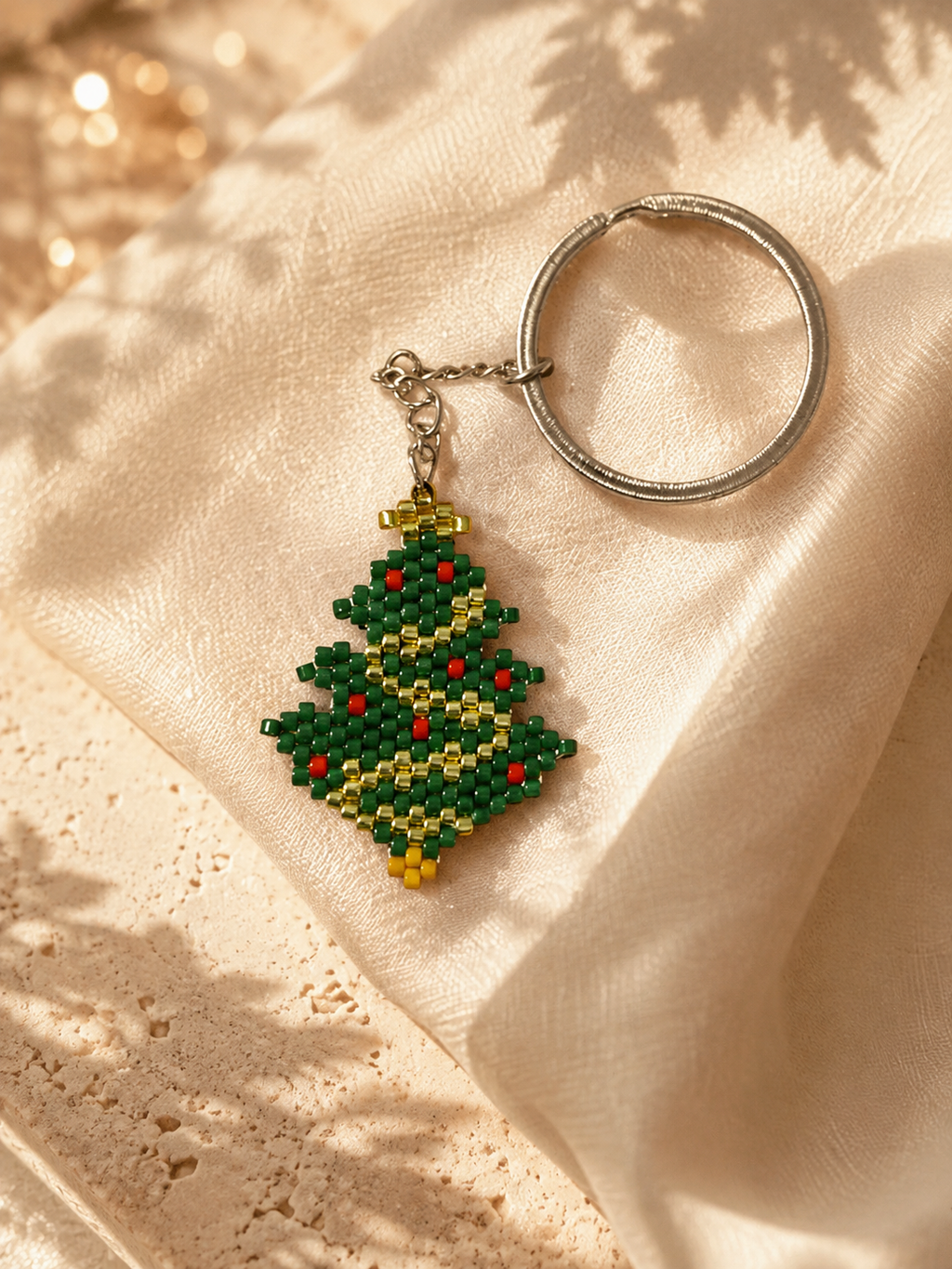 A beaded Christmas tree keychain with a silver metal key ring, resting on draped cream-colored fabric with soft leaf shadows.