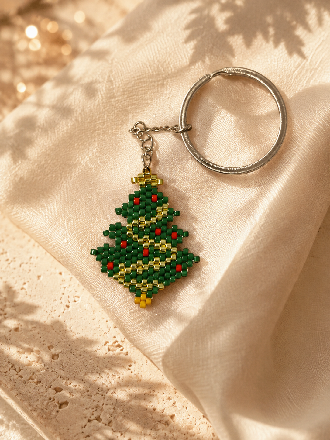 A beaded Christmas tree keychain with a silver metal key ring, resting on draped cream-colored fabric with soft leaf shadows.