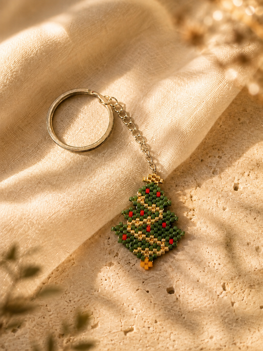 A beaded Christmas tree keychain with a silver-tone split ring and chain, resting on draped beige linen fabric over a textured stone surface.