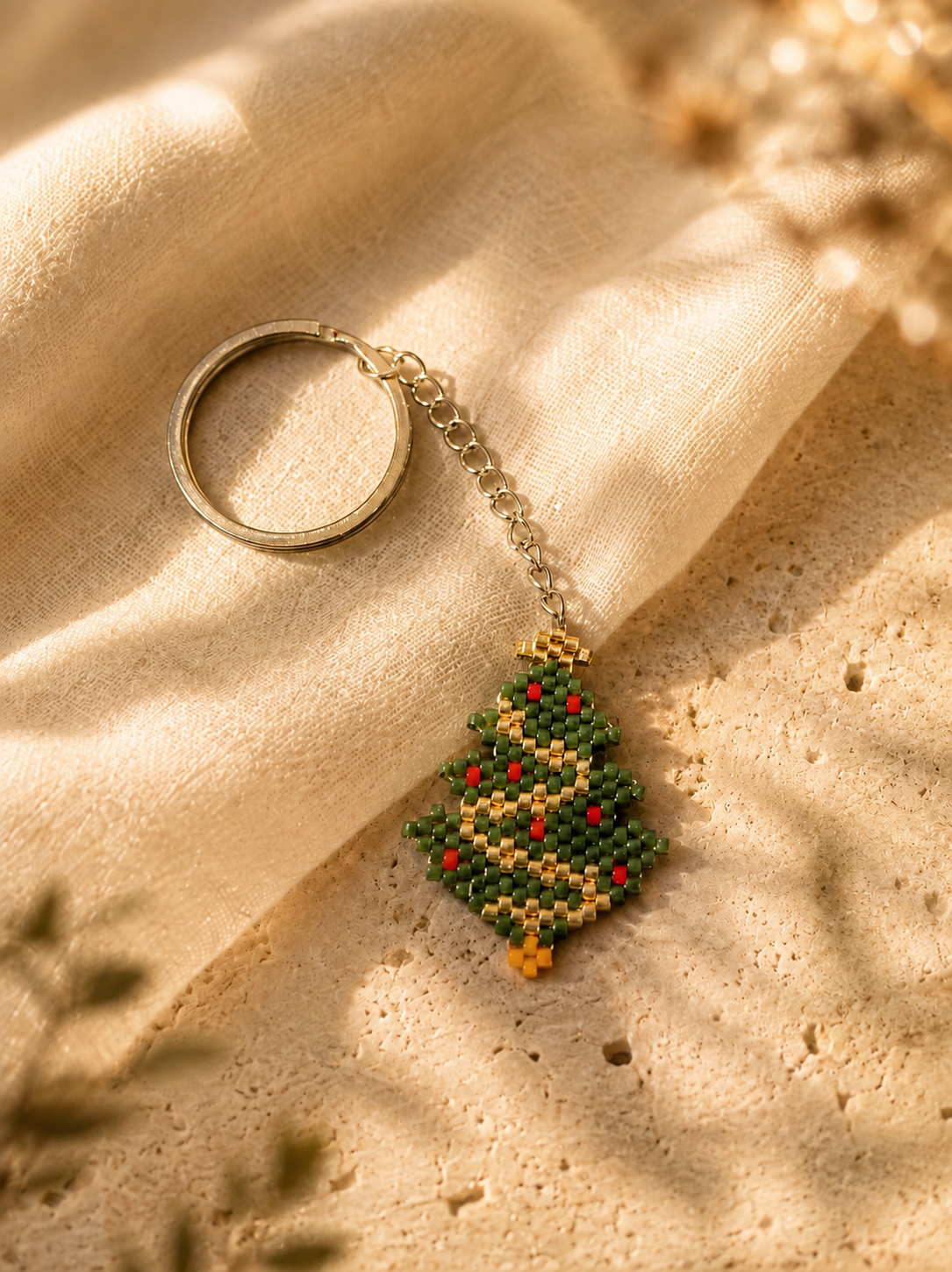A beaded Christmas tree keychain with a silver-tone split ring and chain, resting on draped beige linen fabric over a textured stone surface.