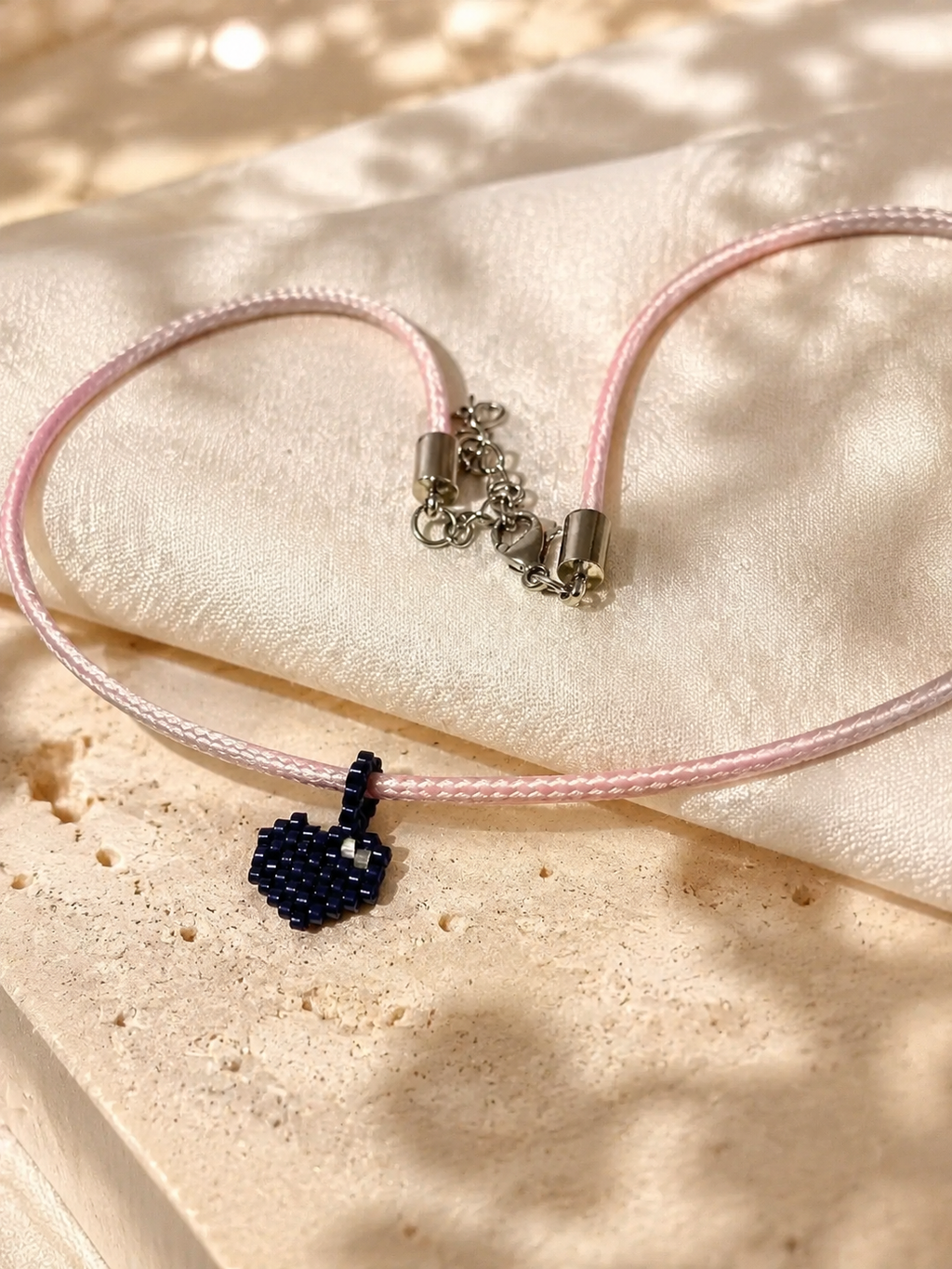 A delicate pink cord bracelet with a small dark blue beaded heart charm, displayed on a textured beige stone surface with soft leaf shadows.