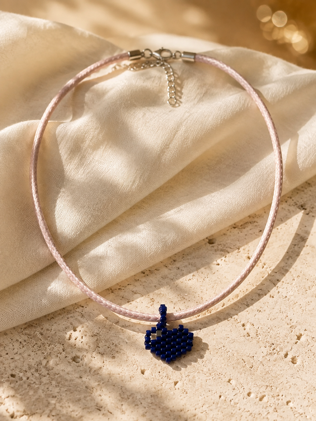 A delicate light pink cord necklace with a small, handcrafted dark blue beaded heart pendant, styled on draped cream fabric over a textured beige stone surface.