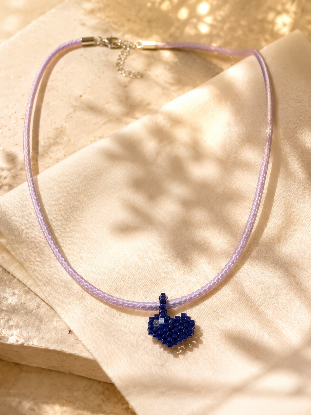 A delicate beaded necklace with a light purple braided cord and a small dark blue beaded heart pendant, displayed on a beige textured surface with soft leaf shadows.
