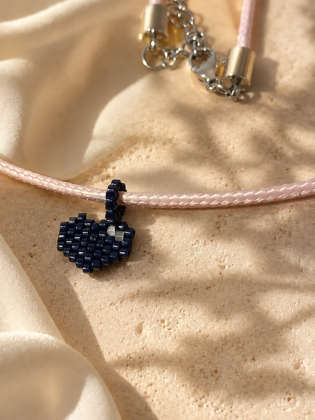 A close-up of a delicate necklace with a light pink braided cord and a small dark blue beaded heart charm, resting on a textured beige surface with soft shadows.
