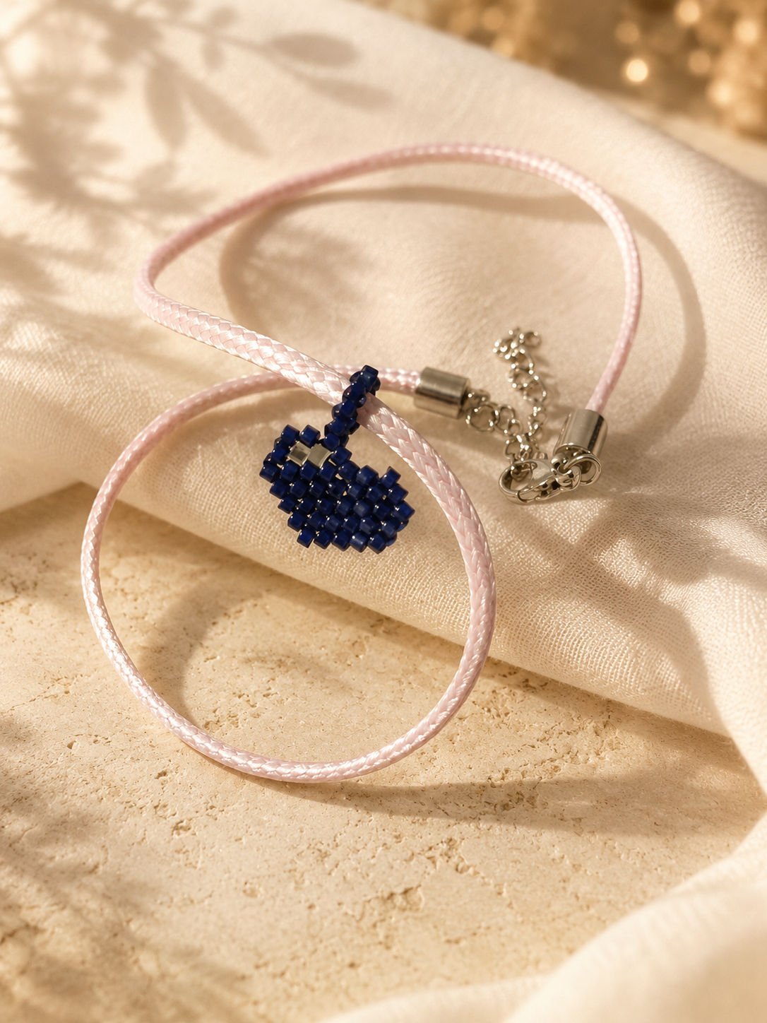 A delicate pink cord bracelet with a small beaded dark blue heart charm and silver-toned metal clasp, displayed on draped beige fabric.