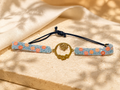 A handmade beaded bracelet with light blue beads, orange floral accents, and a central gold-tone lotus ring, displayed on a beige stone surface with soft fabric in the background.