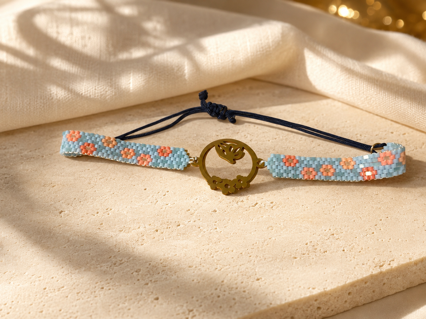 A handmade beaded bracelet with a central gold-tone circular charm, resting on a textured beige stone surface with draped cream fabric in soft shadows.