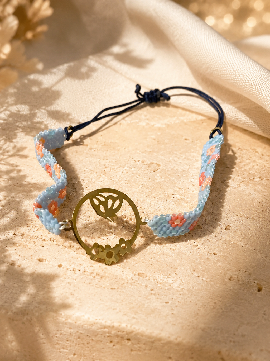 A handmade bohemian-style bracelet featuring a light blue and orange floral woven cord with a central gold-tone circular pendant containing a butterfly and flower motif, resting on a textured beige stone surface with draped cream fabric in the background.