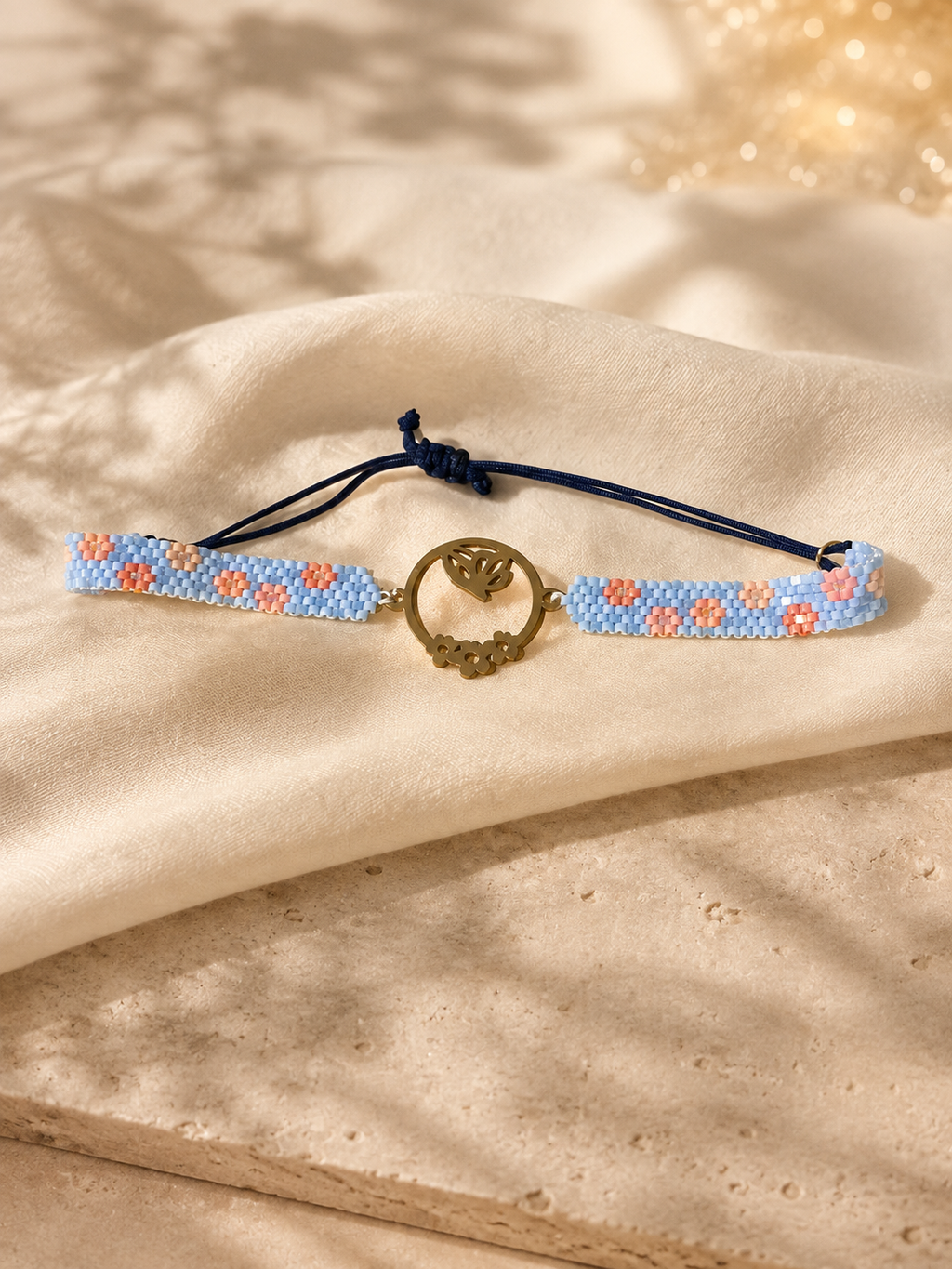 A handmade beaded bracelet with a central gold-tone circular charm, resting on a beige fabric surface with soft shadows.