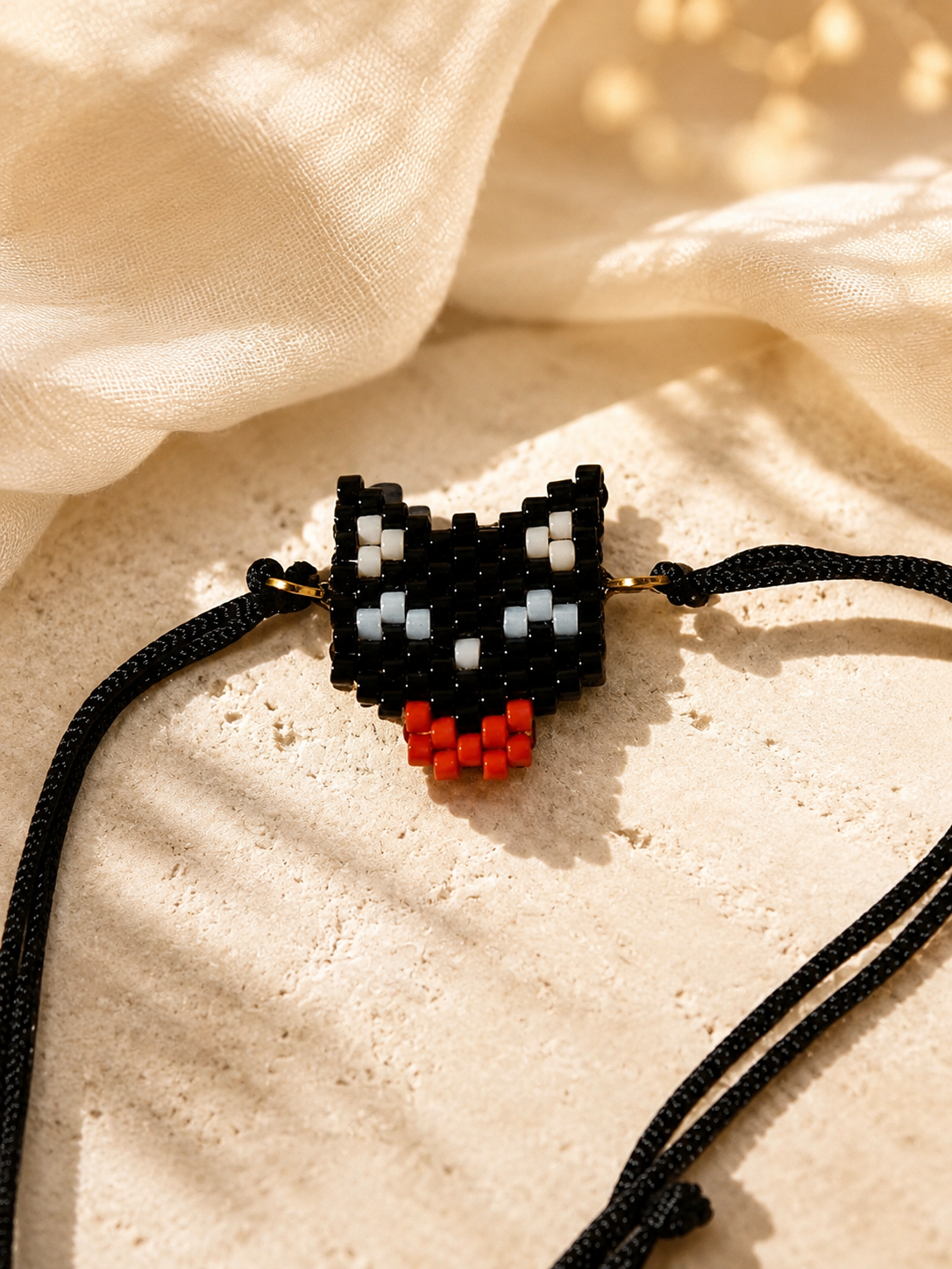 A beaded cat-face pendant necklace on a black cord, styled on a textured beige stone surface with draped cream fabric in warm sunlight.