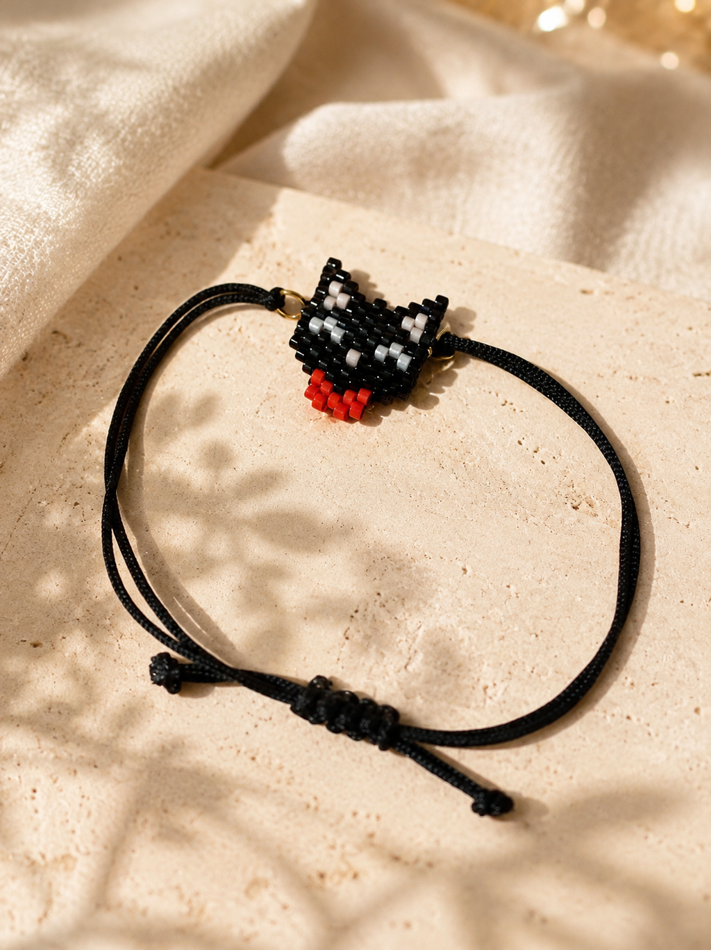 A handmade beaded bracelet with a black cord and a cat-shaped charm featuring black, white, and red beads, styled on a textured beige stone surface with soft fabric in the background.