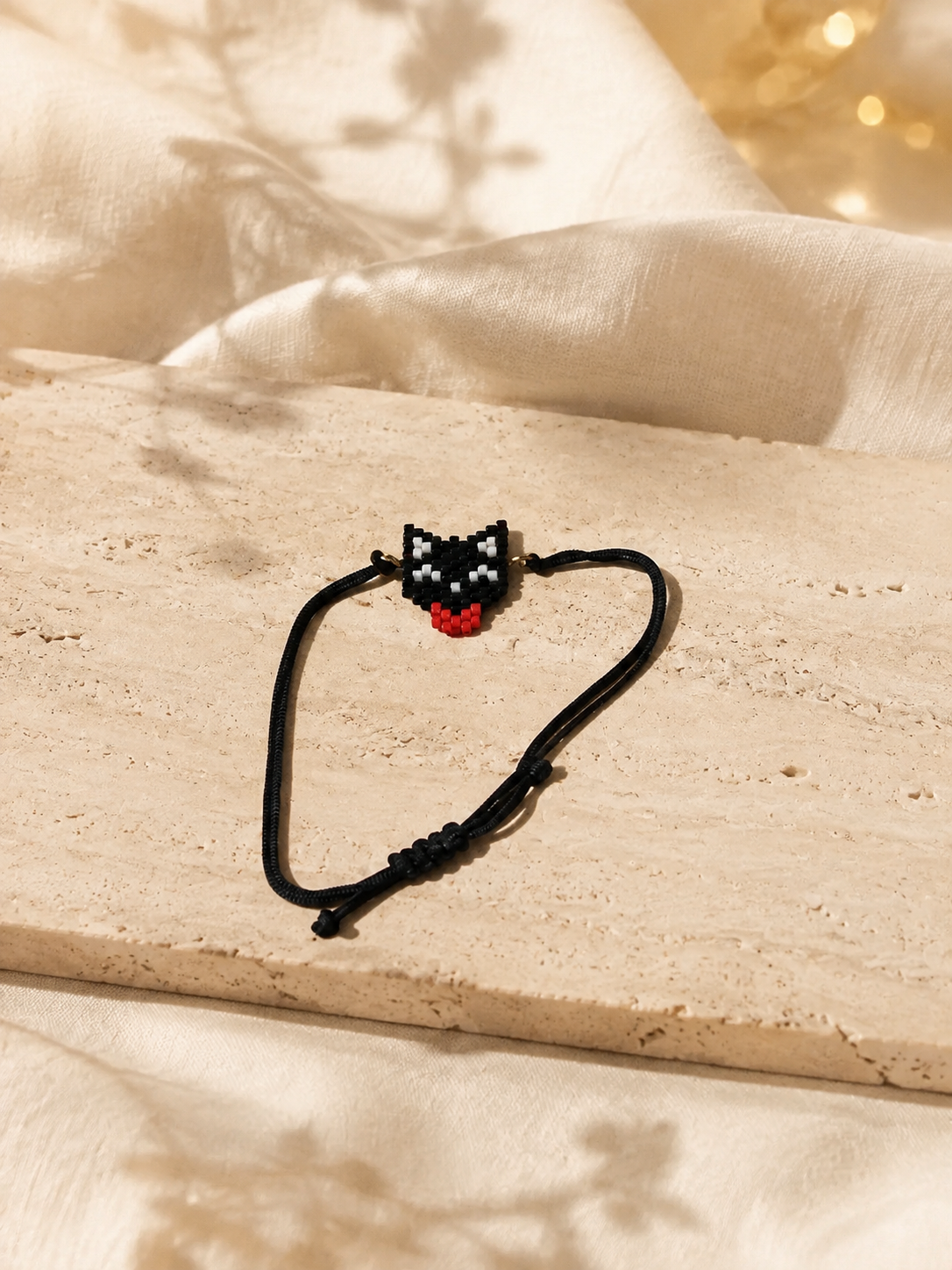 A black adjustable cord bracelet with a small pixel-art cat face charm in black, white, and red, displayed on a beige stone slab with draped cream fabric and warm bokeh lights in the background.