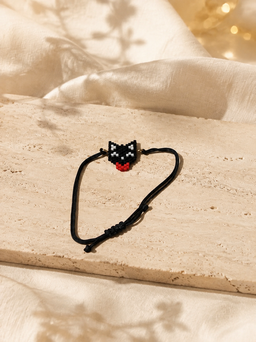 A black adjustable cord bracelet with a small pixel-art cat face charm in black, white, and red, displayed on a beige stone slab with draped cream fabric and warm bokeh lights in the background.