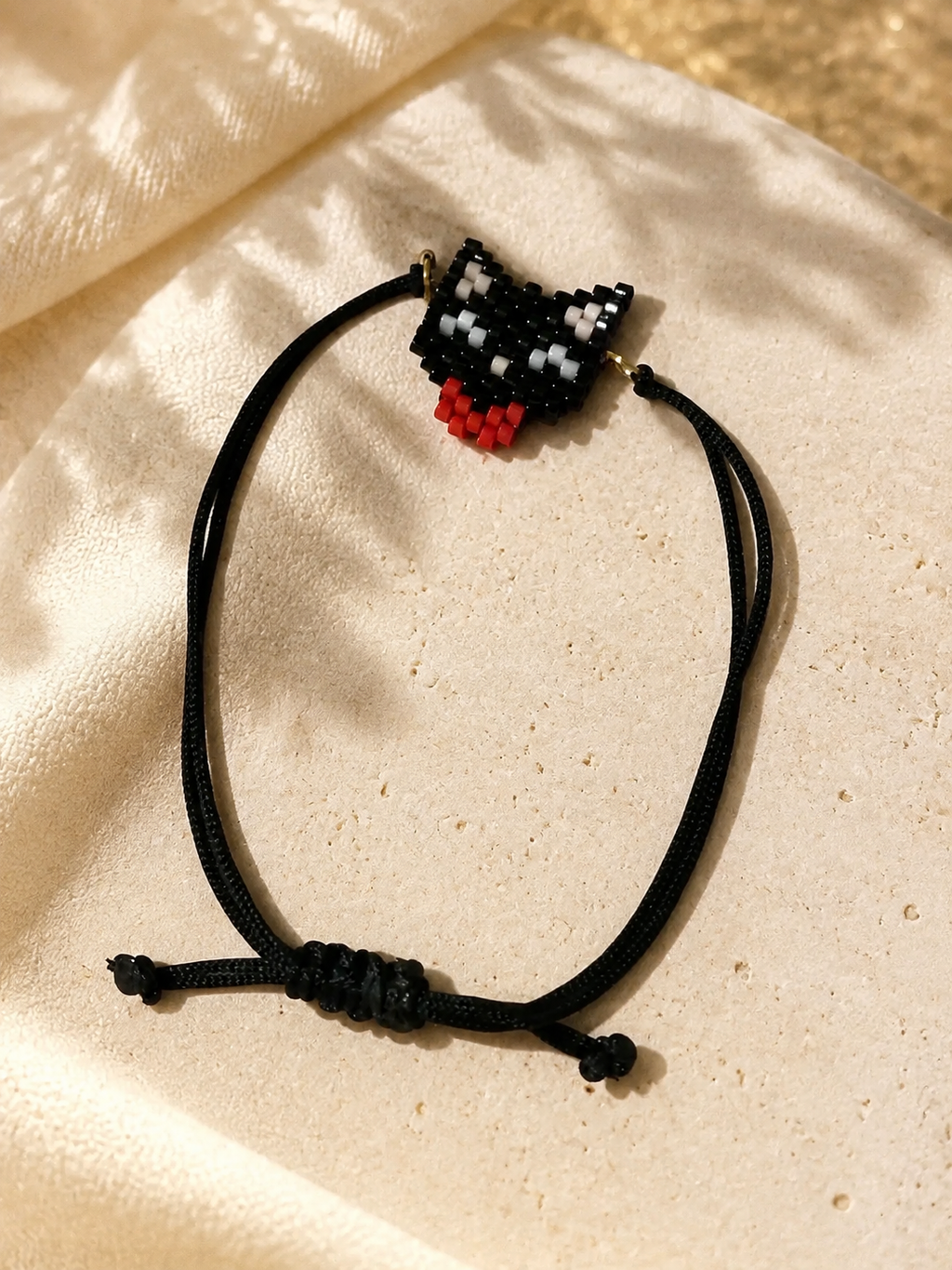 A black adjustable cord bracelet with a small beaded cat face charm in black, white, and red, displayed on a textured beige stone surface with soft fabric shadows.