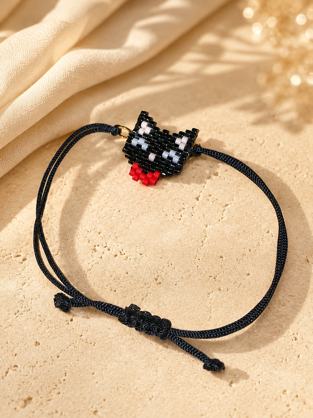 A black cord bracelet featuring a small pixelated black, white, and red cat charm, displayed on a textured beige stone surface with draped cream fabric in the background.