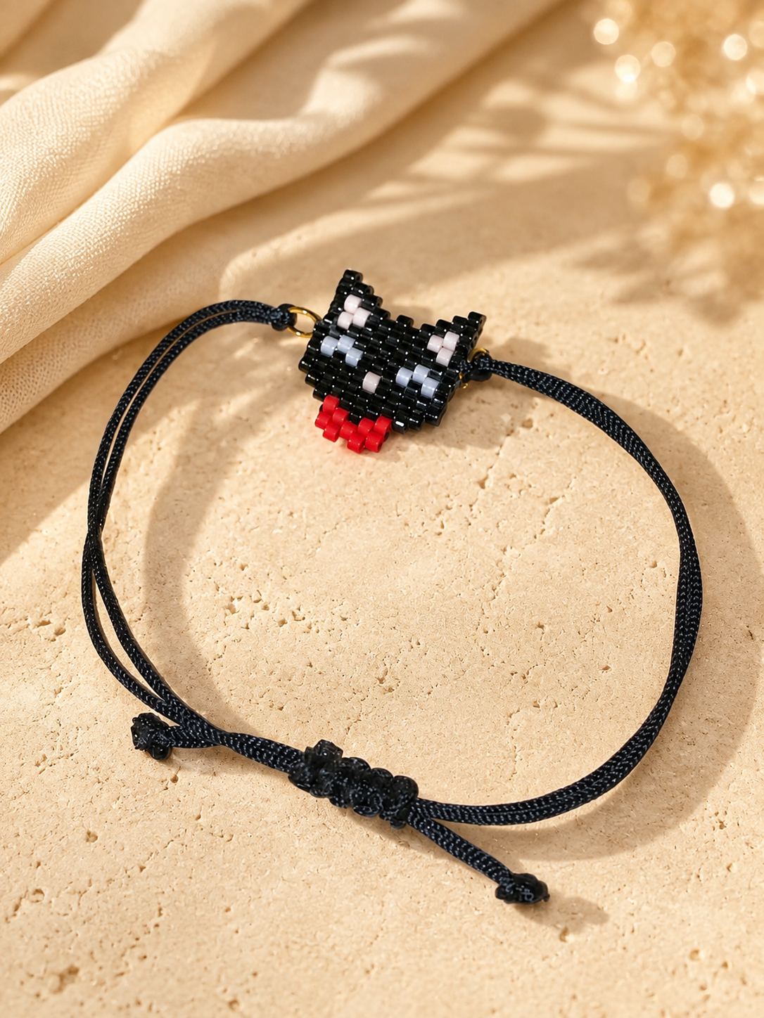 A black cord bracelet featuring a small pixelated black, white, and red cat charm, displayed on a textured beige stone surface with draped cream fabric in the background.