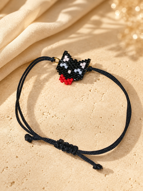 A black cord bracelet featuring a small pixelated black, white, and red cat charm, displayed on a textured beige stone surface with draped cream fabric in the background.