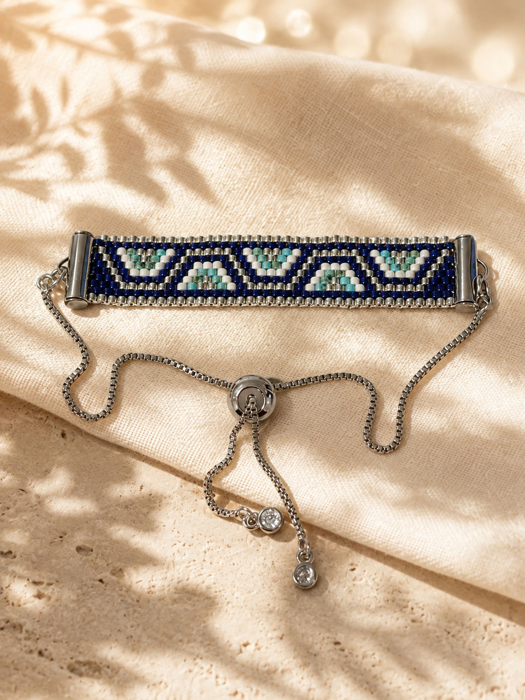 A beaded stretch bracelet with a navy blue and silver geometric pattern, resting on a beige fabric surface with soft leaf shadows.