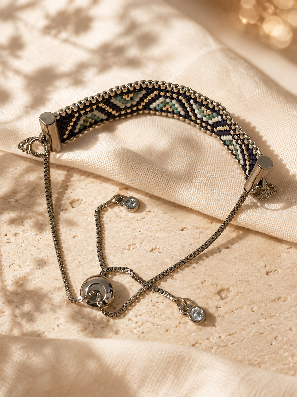 A beaded adjustable bracelet with a geometric pattern in black, gold, and light blue, featuring silver-toned chain extensions and small clear stones, displayed on a textured beige fabric background.