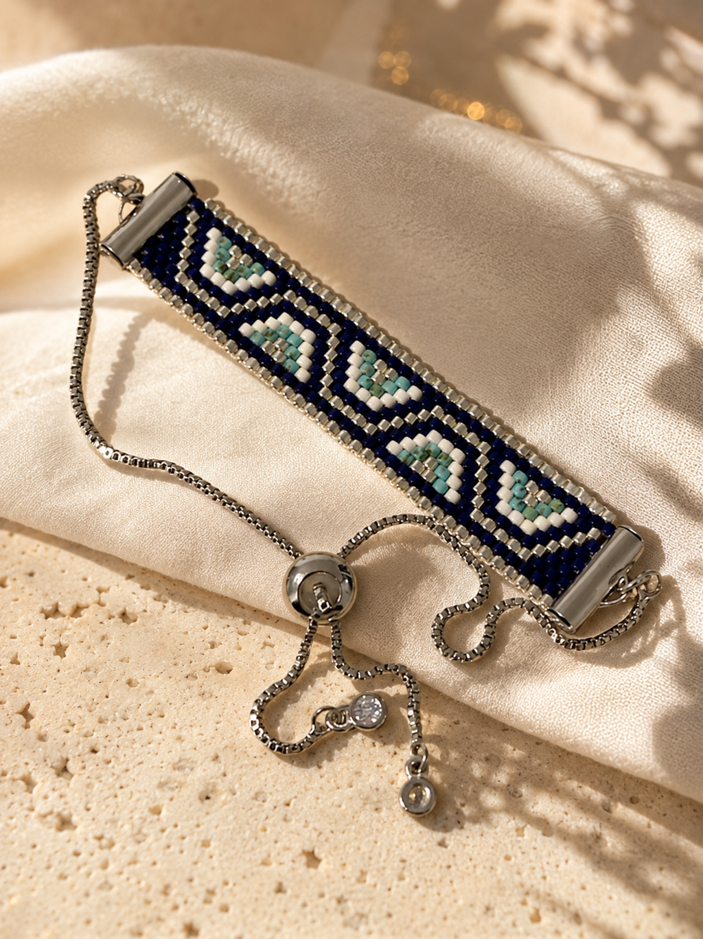 A beaded bracelet with a navy blue and white geometric pattern, accented with mint green and light blue beads, resting on a cream satin fabric over a textured beige surface.