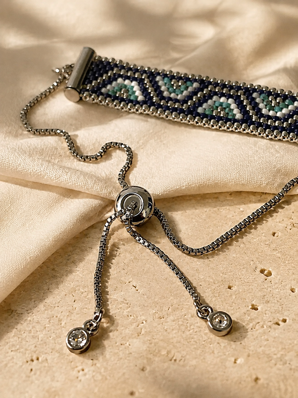 A silver-toned beaded bracelet with geometric patterns in navy, teal, and white, displayed on draped beige fabric alongside a matching silver chain and dangling bezel-set charm.