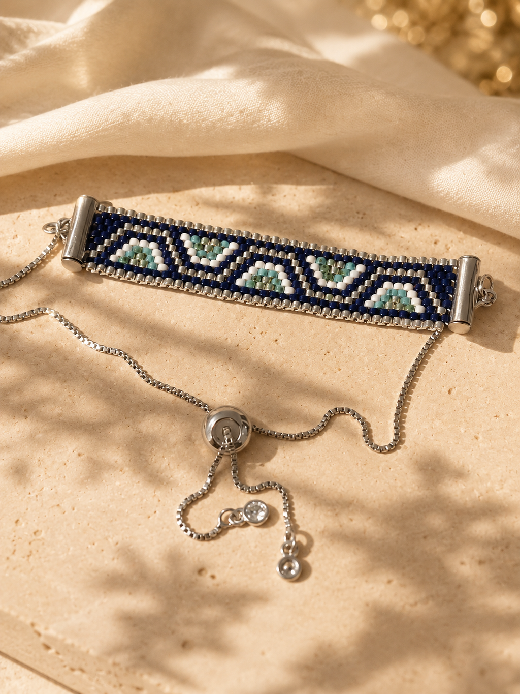 A beaded stretch bracelet with a silver-toned chain and slider clasp, styled on a textured beige stone surface with draped cream fabric and soft shadows.