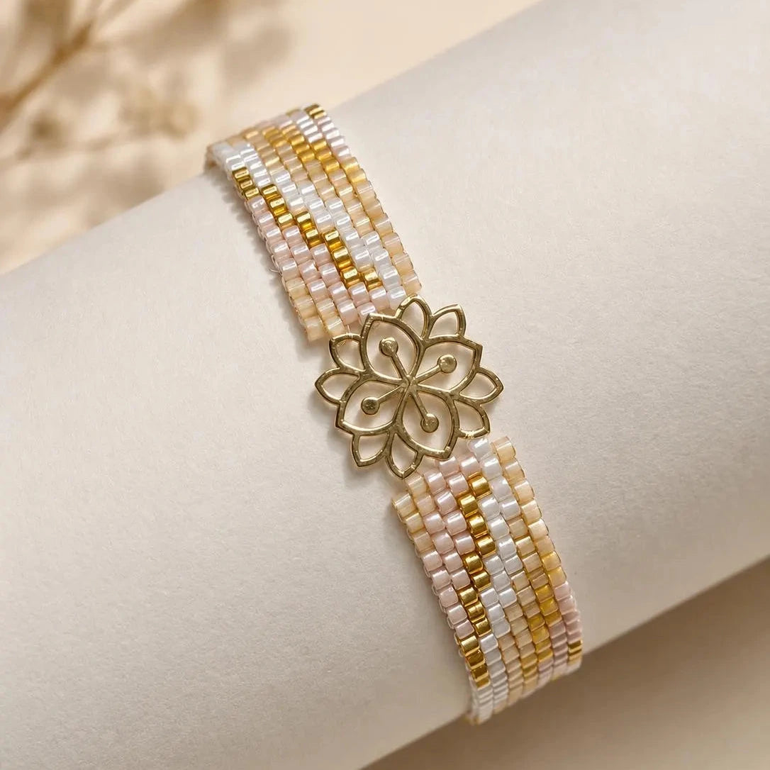 A beaded bracelet with gold and white beads, featuring a central gold flower-shaped charm, displayed on a cream-colored cylindrical stand.