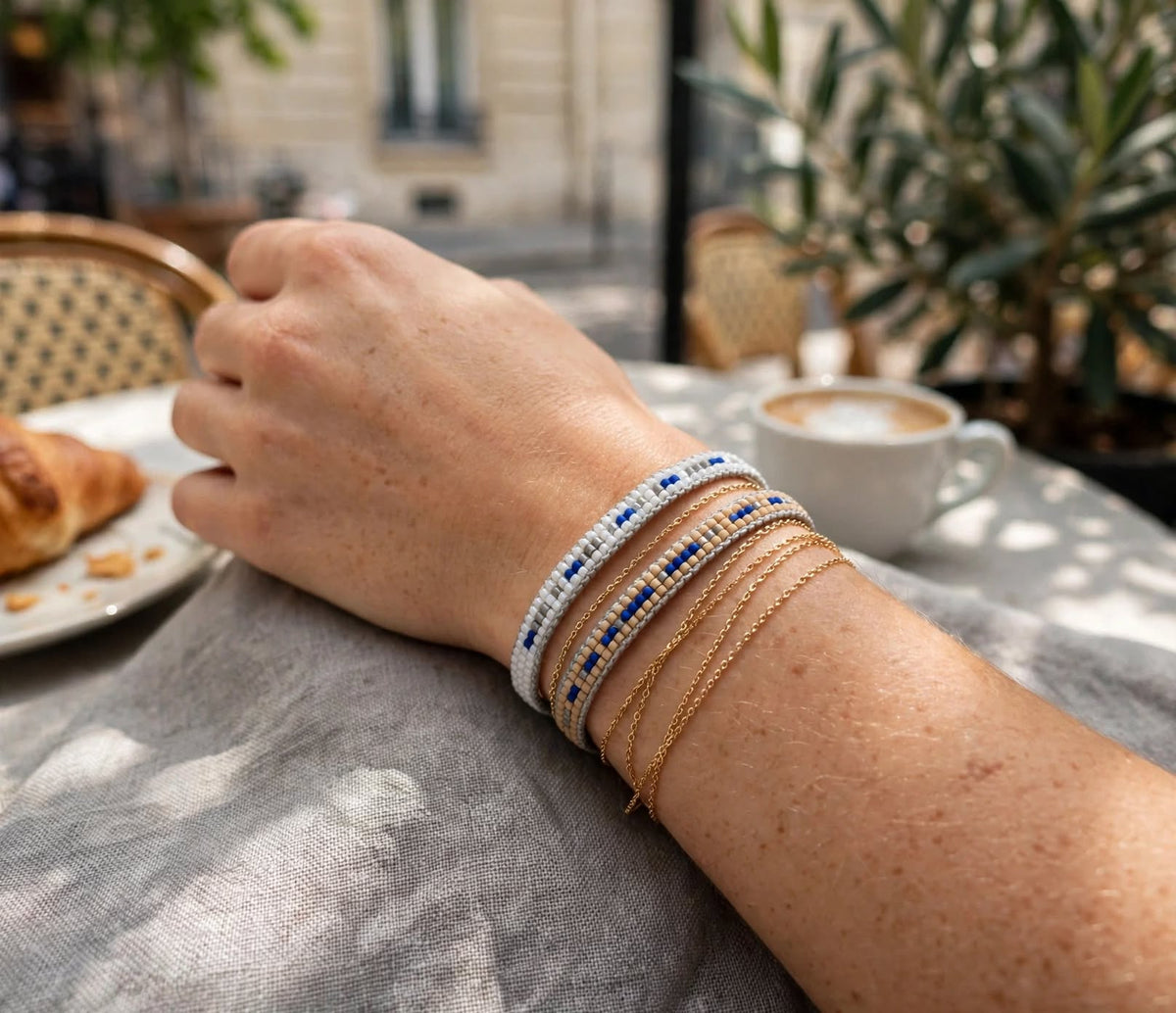 A close-up of a person's forearm resting on a table at an outdoor cafe, wearing several stacked bracelets: a white beaded bracelet with blue accents, a gold chain bracelet, and a gold beaded bracelet. A cup of coffee, a croissant, and plants are blurred in the background.