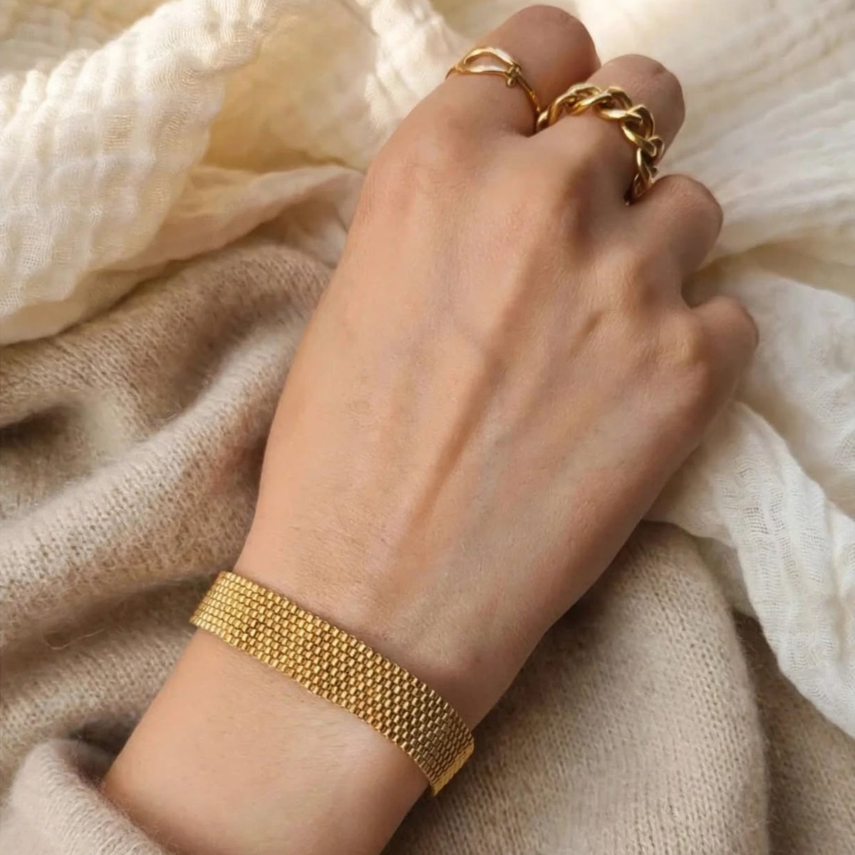 A gold mesh bracelet worn on a person's wrist, with matching gold rings on the fingers, styled against a soft beige knit sweater and cream textured fabric.
