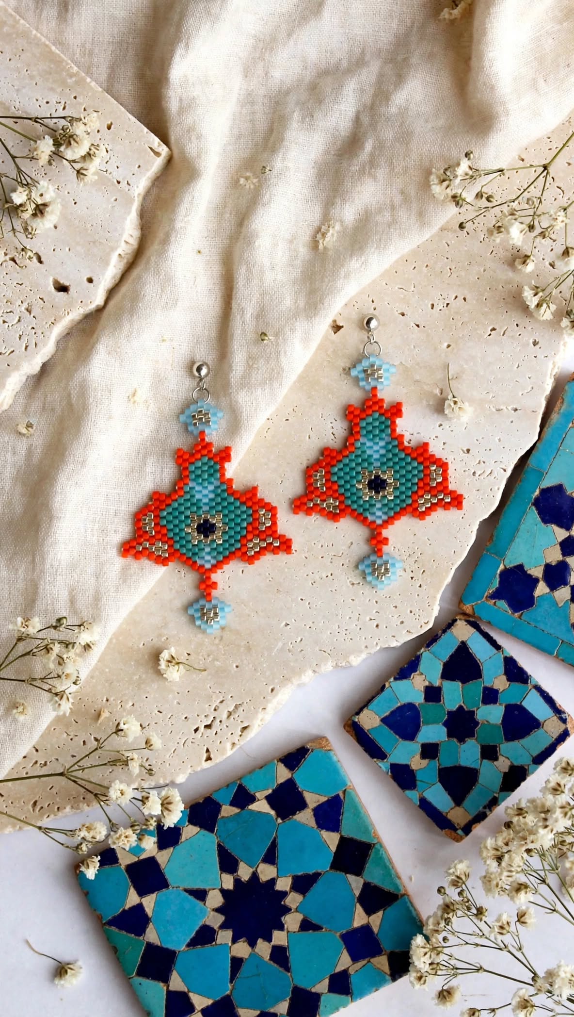 A pair of handmade beaded geometric earrings in turquoise, blue, and orange-red, styled on a beige fabric background with ceramic tiles and dried flowers.