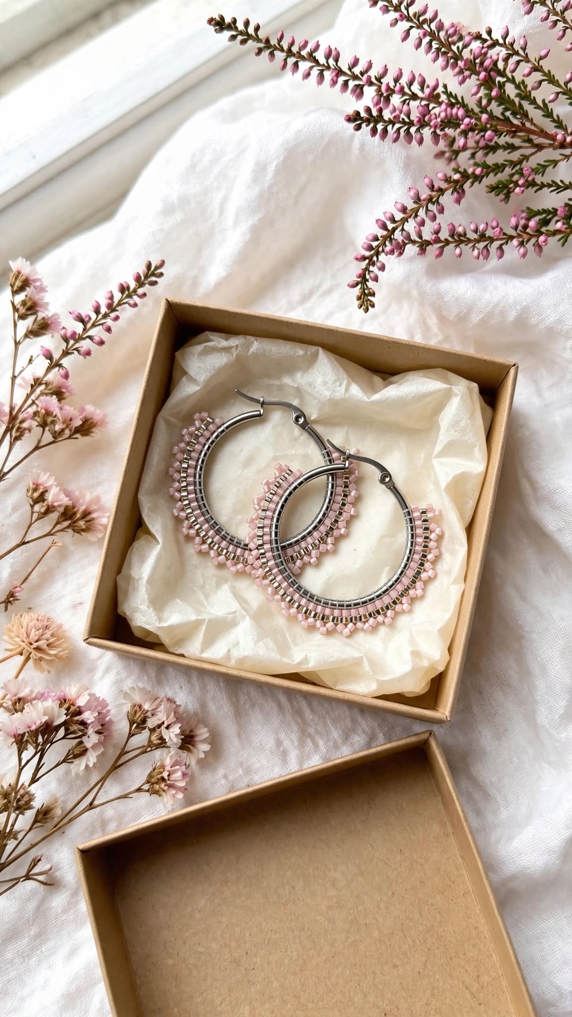 A pair of silver-tone hoop earrings with pink beaded fringe, presented in a brown gift box lined with cream tissue paper, styled on a white fabric background with pink flowers.