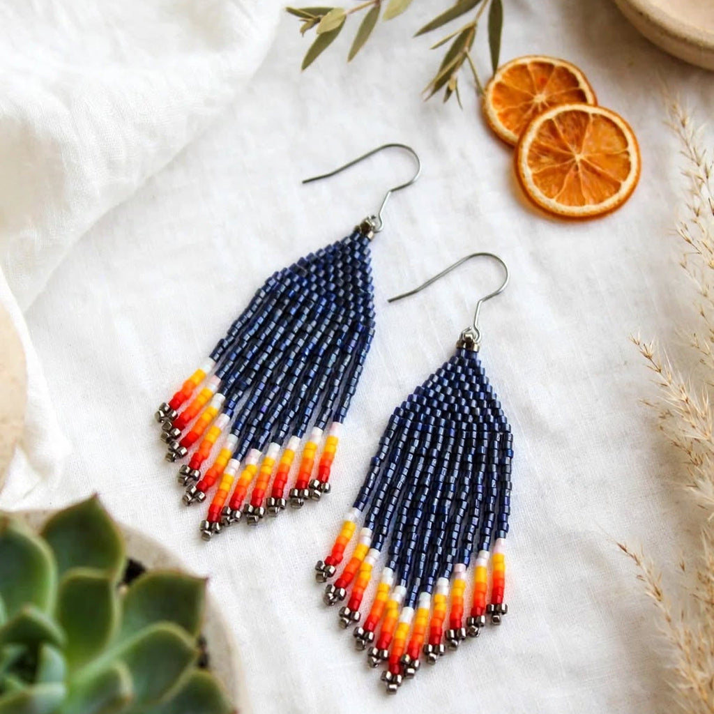 A pair of long beaded dangle earrings with a navy blue top section and a tapered fringe in orange, yellow, and white, displayed on a textured white fabric with dried orange slices, a succulent, and small decorative beads.